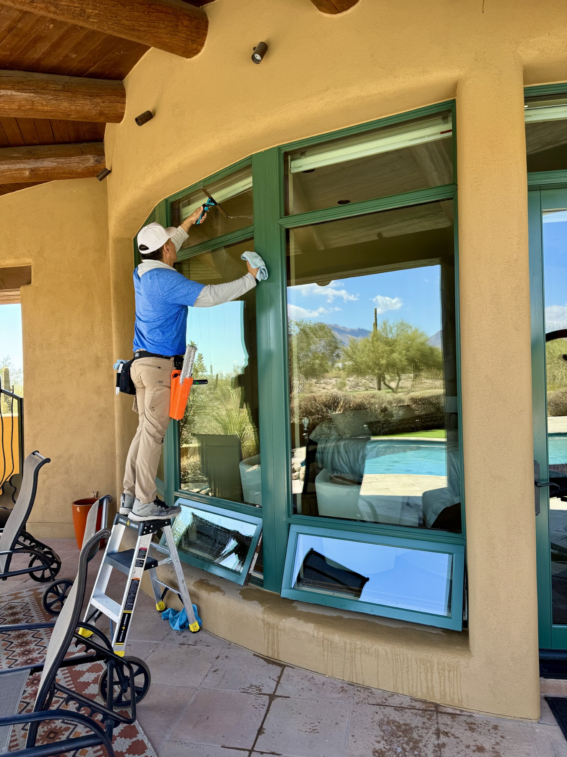 A man cleans patio windows with a squeegee, surrounded by plants and outdoor furniture